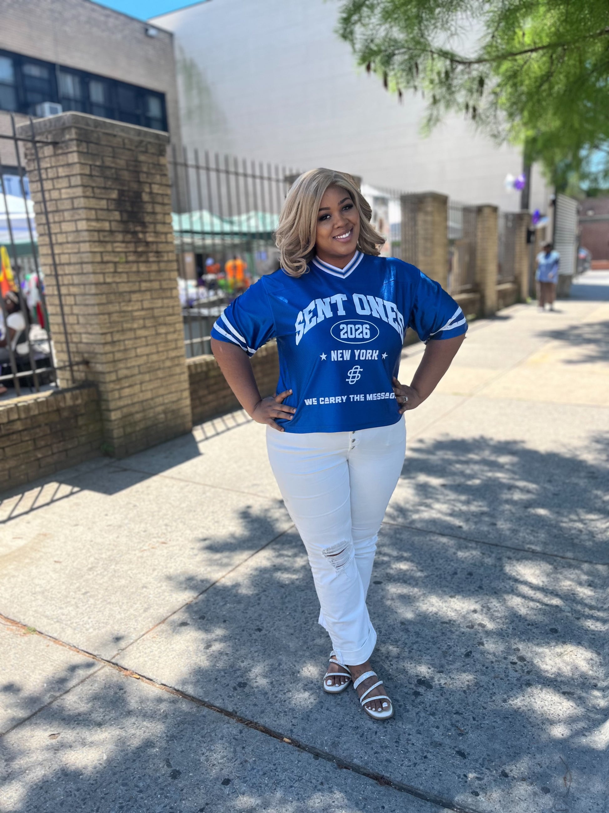 Woman wearing a blue jersey with text standing on a sidewalk.