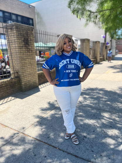 Woman wearing a blue jersey with text standing on a sidewalk.